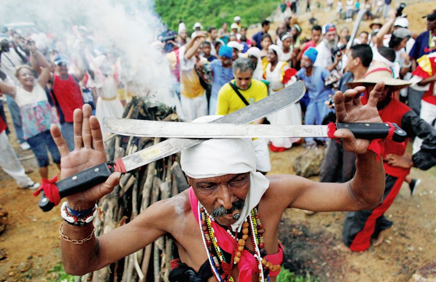 A Santero priest in 2010 holds machetes over his head as he moves around a fire during a Santeria ceremony at the annual Caribbean Festival in Loma del Cimarron, El Cobre, Cuba. (Associated Press) ** FILE **