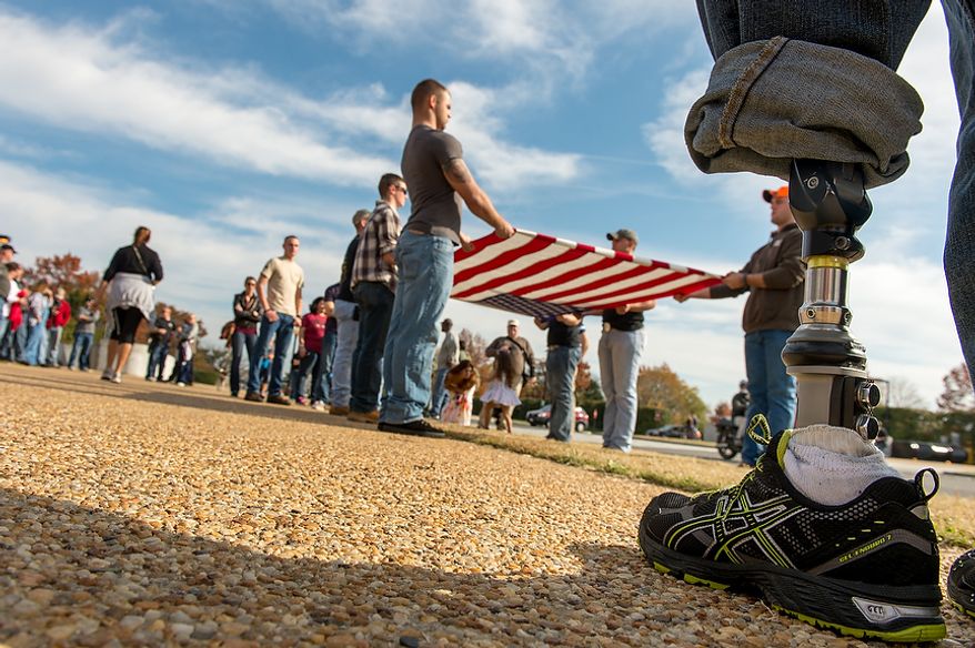 Retired Army Spc. Sean Burke of Lincoln, Neb. who lost his leg from an I.E.D explosion in Afghanistan in 2009, right, watches as off duty men with the Navy Ceremonial Guard, who perform funeral services for service members at Arlington Cemetery, unfold an american flag as veterans and veteran supporters block protesters with the Westboro Baptist Church outside the main entrance to Arlington National Cemetery on Veterans Day, Arlington, Va., Monday, November 12, 2012. Approximately a hundred veterans and veteran supporters came out to block the protest by the Westboro Baptist Church, based in Topeka, Kan., which is known for extreme views and have caused an uproar over the past few years for their protests. (Andrew Harnik/The Washington Times)