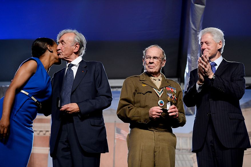 Former President Bill Clinton (right) and Scottie Ooton (second from right), a member of the 84th Infantry Division, which liberated the Hanover-Ahlem Concentration Camp in Germany in World War II, applaud as U.S. Holocaust Memorial Museum staff member Rebecca Dupas (left) gets a kiss from Elie Wiesel, founding chairman of the museum. He and Mr. Ooton were presented with commemoration pins during the U.S. Holocaust Memorial Museum's 20th-anniversary commemoration and national tribute to Holocaust survivors and World War II veterans in Washington on Monday, April 29, 2013. (Andrew Harnik/The Washington Times)