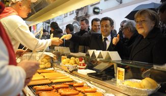 **FILE** French President Nicolas Sarkozy (center) and German Chancellor Angela Merkel (right) stop at a roast sausage fast food stand as they visit the Christmas market in Freiburg, Germany, on Dec.  10, 2010. (Associated Press)