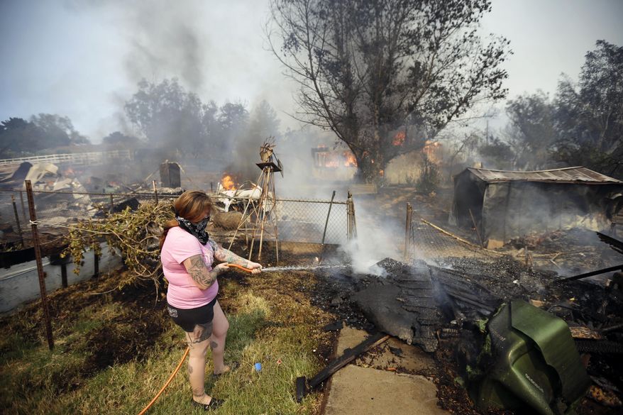A woman douses water from a hose around her home as her neighbor&#x27;s home burns during a wildfire Thursday, May 15, 2014, in Escondido, Calif. One of the nine fires burning in San Diego County suddenly flared Thursday afternoon and burned close to homes, trigging thousands of new evacuation orders. (AP Photo/Gregory Bull)