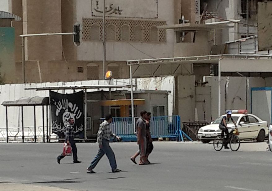 This image taken with a mobile phone camera shows people passing a black flag used by Islamic militants and a police cruiser belonging to the Islamic State group in front of the provincial government headquarters in Mosul, 225 miles (360 kilometers) northwest of Baghdad, Iraq, Tuesday, July 15, 2014. The panic that initially gripped Iraq after Sunni militants led by the Islamic State extremist group seized the country's second-largest city, Mosul, and swept across northern and western Iraq has largely subsided.(AP Photo)