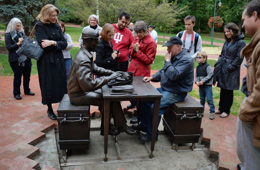 In this photo taken on Thursday, Oct. 9, 2014, artist Tuck Langland demonstrates how he envisioned visitors could sit at the desk with Ernie Pyle during the installation of the bronze Ernie Pyle statute outside Franklin Hall at Indiana University in Bloomington, Ind. The statue depicts Pyle sitting on an ammunition box with his typewriter, notes and a coffee cup on a table. (AP Photo/The Herald-Times, Chris Howell)
