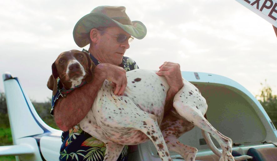 In this undated photo provided by National Geographic Channels, veterinarian Dr. Scott Sims, the star of Nat Geo Wild's new "Aloha Vet" series, carries a dog next to the two-seater airplane he built from a kit, near his facility at Kilauea on the island of Kauai in Hawaii. The series follows Sims on his rounds, from his clinic on Kauai to patients on other islands. His patients really do cover all creatures great and small, from dogs to horses and birds to sea turtles. The 59-year-old Renaissance man figures he’s treated over 130,000 animals since setting up practice in 1984 in Novato, Calif. He moved his practice to Hawaii in 2001. (AP Photo/National Geographic Channel)