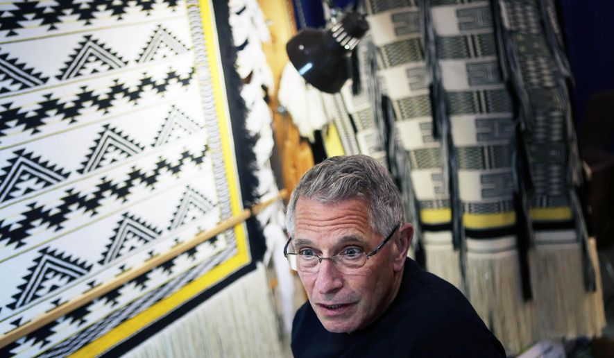In this Aug. 2014 photo, Salem native John Beard stands before his Ravenstail weaving display at the 2014 Oregon State Fair in Salem, Ore.. Salem native John Beard of Tigard has been making connections at the Oregon State Fair for more than three decades. He’s made connections between warp and weft, the vertical and horizontal yarns that he weaves on his loom, but demonstrating the art of Ravenstail weaving is about more than the large and beautiful ceremonial robes he produces. (Thomas Patterson/Statesman-Journal via AP) MANDATORY CREDIT