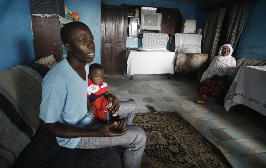 In this photo taken Tuesday, Aug. 18, 2015, nurse Donnell Tholley, 25, holds his adopted son Donnell Junior at their apartment in Freetown, Sierra Leone, as the baby’s great-grandmother Marie looks on. Tholley adopted the boy following the death of the mother Fatu Turay last year from the Ebola virus at the hospital on the outskirts of Freetown, Sierra Leone. The Ebola epidemic killed nearly 4,000 Sierra Leoneans and left thousands of orphans. (AP Photo/Sunday Alamba)