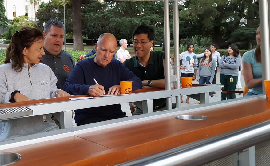 In this photo provided by the California Governor's Press Office, California Gov. Jerry Brown signs a bill allowing beer bikes to operate on city streets as his wife Anne Gust Brown, left, looks on Sunday, Oct. 4, 2015, in Sacramento, Calif. Brown signed the bill before going on a ride with his wife and Sen. Richard Pan, a Sacramento Democrat who crafted the bill to create a vehicle safety standard for bike buses that allow up to 15 people to pedal at once. (California Governor's Press Office via AP) MANDATORY CREDIT