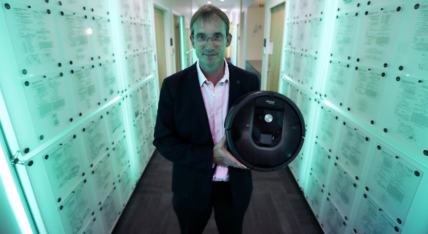 In this Thursday, Aug. 25, 2016, photo, iRobot co-founder and CEO Colin Angle is illuminated in blue-green light while holding a Roomba vacuum in a hallway decorated in patents the company owns, at their headquarters in Bedford, Mass. Angle is transitioning the company from military projects to household robots. (AP Photo/Charles Krupa)