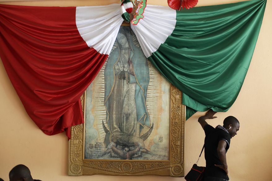 In this Sept. 26, 2016 photo, a Haitian man listening to music on his phone and leaning against a wall decorated with a Mexican national flag and a framed image of the Virgin of Guadalupe, waits for a bed at the Padre Chava migrant shelter in Tijuana, Mexico. Many Haitians arriving at the U.S. border with Mexico are unaware of a new U.S. policy of putting them in deportation proceedings and detaining them while making efforts to fly them home. (AP Photo/Gregory Bull)