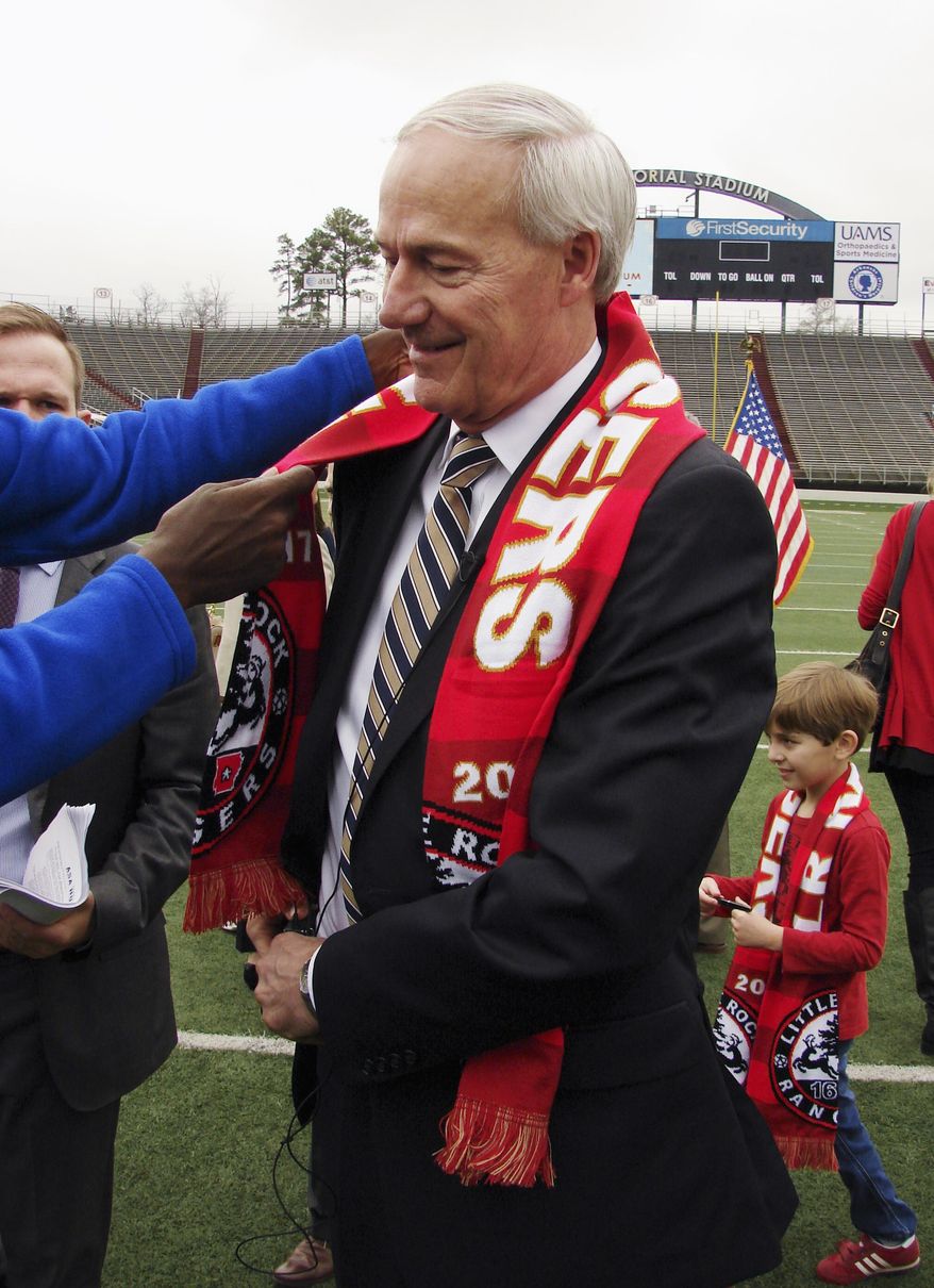 A television cameraman adjusts a scarf as he reaches in to place a microphone on Gov. Asa Hutchinson as the governor prepares to talk to reporters Wednesday, Feb. 22, 2017, about bills in the Arkansas Legislature that would impose taxes on online purchases. Hutchinson, who had just been given a scarf from the Little Rock Rangers soccer team, spoke on the floor of War Memorial Stadium in Little Rock. (AP Photo/Kelly P. Kissel)