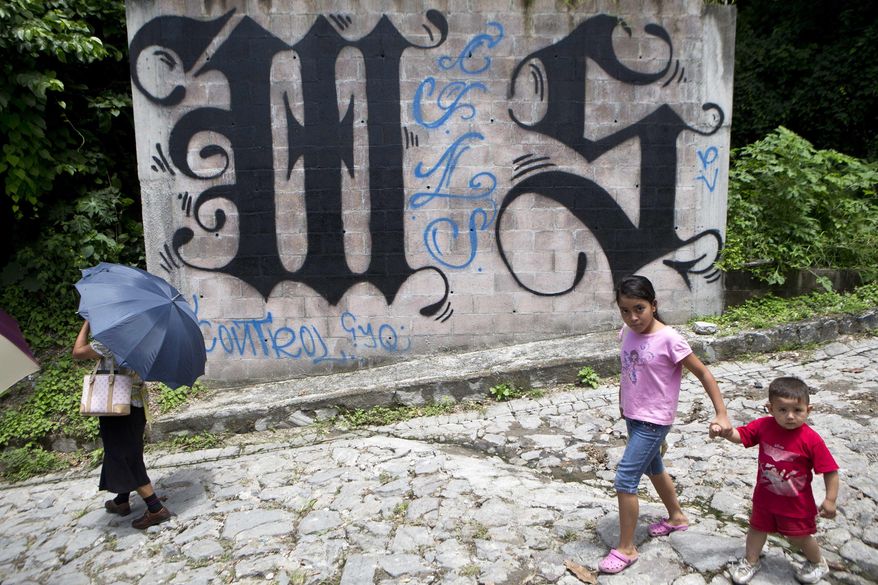 A family walks by a wall covered by a symbol from the Mara Salvatrucha, of MS-13 gang in Ilopango, El Salvador, on Aug. 21, 2014. In communities across El Salvador, the Mara Salvatrucha and their arch-rivals, the 18th Street Gang, are de facto rulers. (AP Photo/Esteban Felix) **FILE**