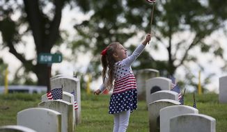 Four-year-old Grace Hendrian, of Quincy, Ill., stops to play with a small American flag while helping set the flags in front of tombstones in Sunset Cemetery Wednesday, May 24, 2017, on the grounds of the Illinois Veterans Home in Quincy, Ill. Scores of volunteers turned out to help place the flags in honor of Memorial Day. (Phil Carlson/Quincy Herald-Whig via AP)