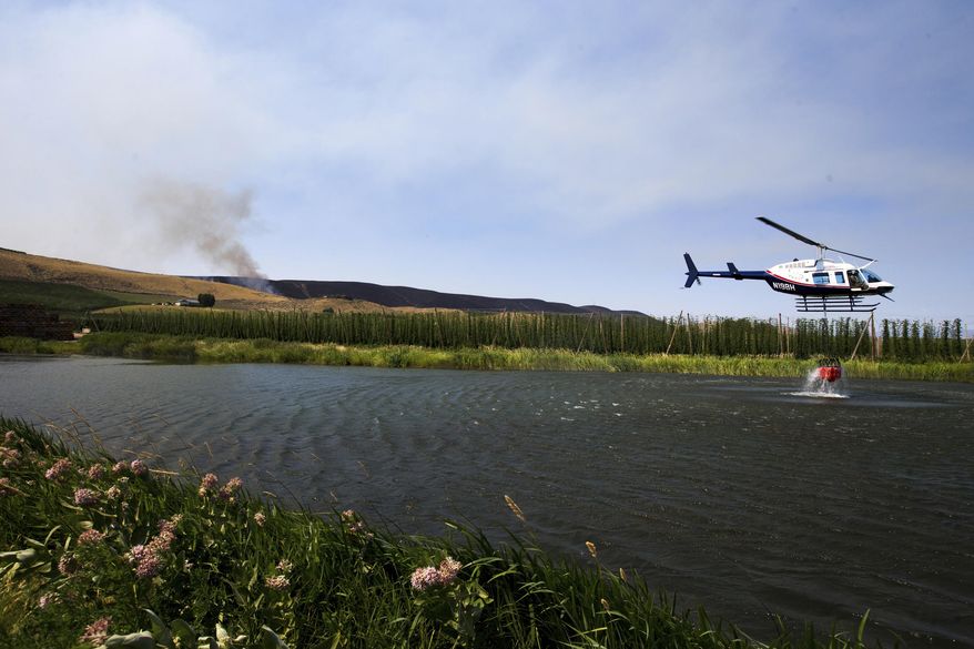 As Rattlesnake Ridge continues to burn in the distance, a fire fighting helicopter loads water into a bucket in Moxee, Wash., on Thursday, July 6, 2017. The fire burned more than 5,500 acres of mostly grass and brush in less than 24 hours, and threatened about 30 homes. (Jake Parrish/Yakima Herald-Republic via AP)