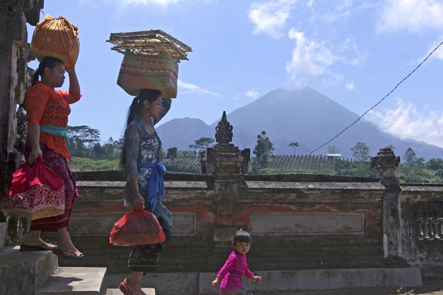 Women carry offerings at a temple about 10 kilometers (6.3 miles) from Mount Agung volcano, rear, at Pemuteran village in Karangasem, Bali, Indonesia, Wednesday, Sept. 27, 2017. Warnings that a volcano on the Indonesian tourist island of Bali will erupt have sparked an exodus of more than 75,000 people that is likely to continue to swell, the country's disaster agency said. (AP Photo/Firdia Lisnawati)