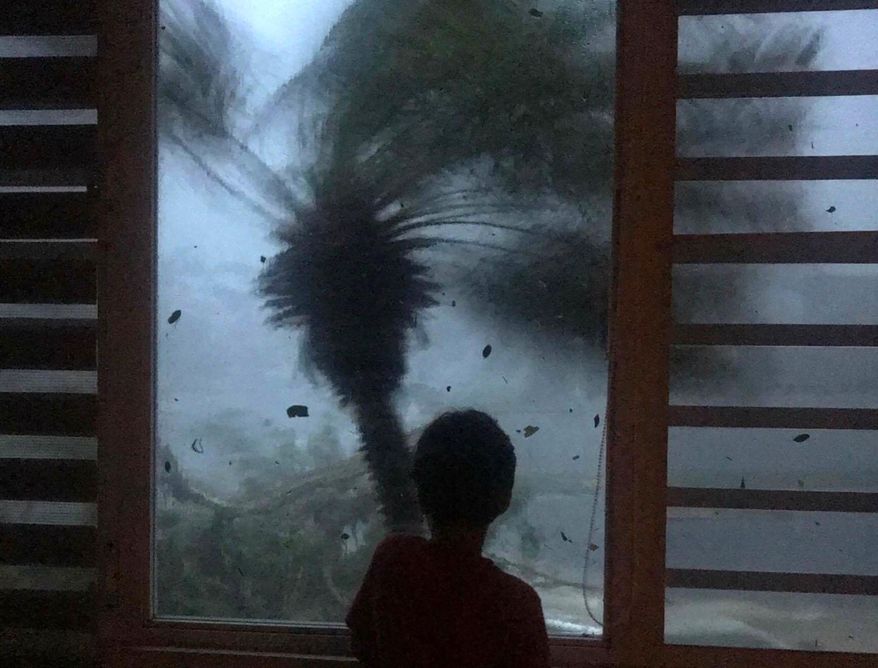 In this early morning Sept. 20, 2017 photo, a young boy looks out the window as strong winds brought on by Hurricane Maria bend a palm tree and send debris flying, in Juncos, Puerto Rico. As rains began to lash Puerto Rico, Gov. Ricardo Rossello warned that Maria could hit "with a force and violence that we haven't seen for several generations." (AP Photo/Linda Rodriguez Flecha)