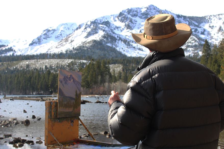 In a Monday, Jan. 22, 2018 photo, artist Charles Muench paints the view of Mount Tallac from a beach at Fallen Leaf Lake in South Lake Tahoe, Calif., creating a piece he calls “Clouds Clearing – Mt. Tallac.” (Claire Cudahy/The Tahoe Tribune via AP)