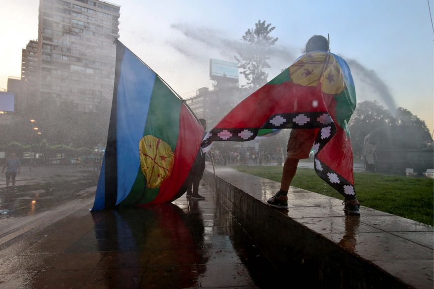 Protesters with Mapuche flags watch a police a water cannon disperse demonstrators during a protest commemorating the ten year anniversary of the police killing of Mapuche indigenous activist Matias Catrileo, Santiago, Chile, Friday, Jan. 5, 2018. Catrileo was shot to death on Jan. 3, 2008 by a police officer during a land dispute in southern Chile. The officer was sentenced to three years in jail, but served his time on probation. The officer was eventually removed from the police force. (AP Photo/Esteban Felix)