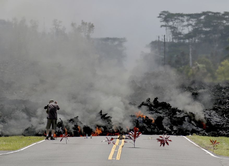 Lava burns across a road in the Leilani Estates subdivision as an unidentified person takes pictures of the flow, Saturday, May 5, 2018 near Pahoa, Hawaii. Offerings of Hawaiian ti leaves, rocks and cans to the fire goddess Pele lie in the street in front of the lava. (AP Photo/Caleb Jones)