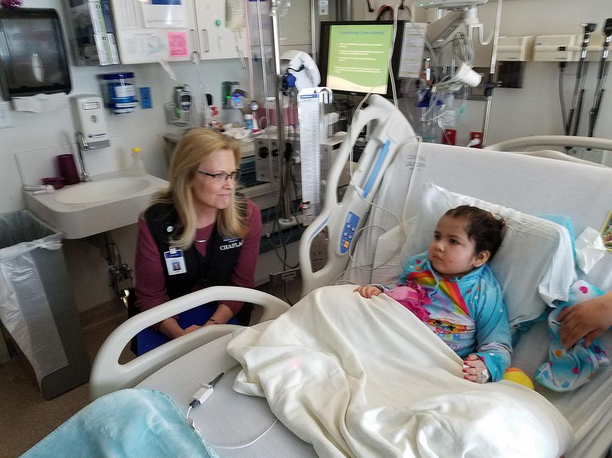 In this April 12, 2018, photo, Chaplain Vicki Pond, left, sits with Ruthie Yanez, 5, at Primary Children's Hospital in Salt Lake City. Surgery to remove a brain tumor has left Ruthie’s soft, chocolate brown eyes sightless; her overall condition remains “guarded” after several weeks at the hospital. Still, her face brightens into a smile upon hearing Pond pull up a chair next to her bed. (Bob Mims/The Salt Lake Tribune via AP)