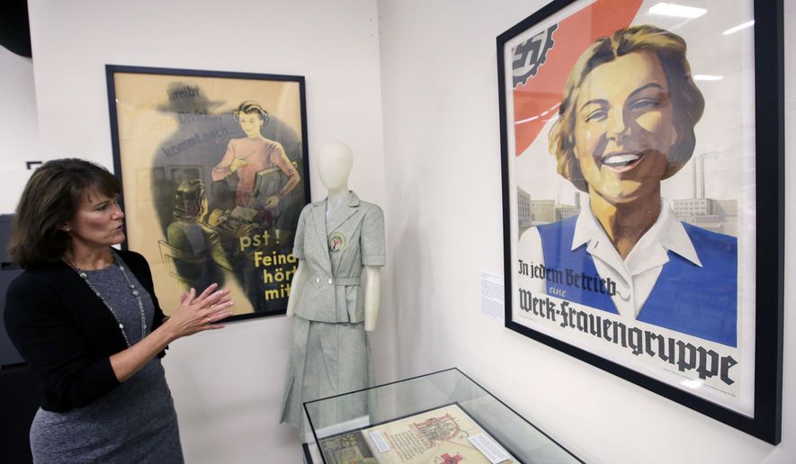 In this Monday, May 21, 2018, photo, Sue Wilkins, director of education at The International Museum of World War II, in Natick, Mass., stands near a 1933 propaganda poster, right, that praised the Nazi organization German Labor Front, which was created after the Nazis eliminated trade unions. A mannequin, center, displays a uniform of the Nazi Lebensborn program, designed to be worn by women bearing children considered by the state as racially valuable. They are part of a new exhibition called &amp;quot;Women in WWII: On the Home Fronts and the Battlefronts&amp;quot; at the museum explores the important and unconventional roles women played in every large nation that fought in World War II. (AP Photo/Steven Senne)
