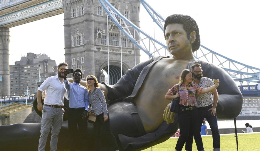 People take photos by a 25ft statue of actor Jeff Goldblum in a pose from a scene in the first Jurassic Park movie, which has been created by a TV channel to celebrate the film&#39;s 25th birthday, at Potters Fields Park, London, Wednesday July 18, 2018. Tower Bridge in the background.  (Doug Peters/PA via AP)
