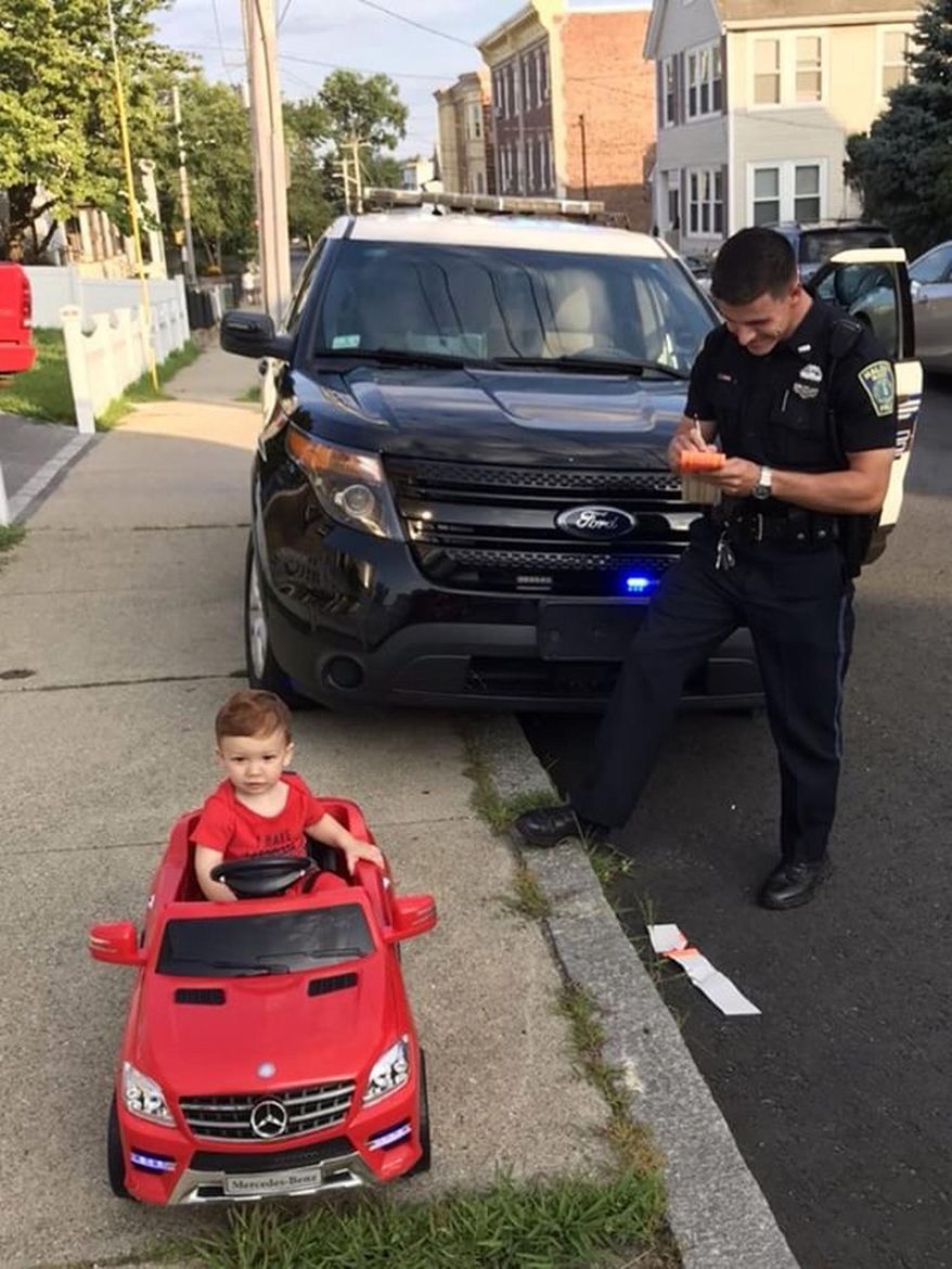 In this July 31, 2018 photo, Grayson Salerno, 1, is pulled over for driving without a license, but he got off with a "cuteness warning" in Malden, Mass. Police in the Boston suburb said they staged the traffic stop after spotting Salerno driving a red toy Mercedes convertible along a sidewalk. (Cori-Devan Salerno via AP)