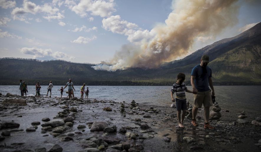 FILE - In this Sunday, Aug. 12, 2018 file photo provided by the National Park Service, people walk along the shore near Lake McDonald Lodge as the Howe Ridge Fire burns in Glacier National Park, Mont. Wildfires that have kept portions of Glacier National Park closed for two weeks are scrambling visitors’ plans and prompting some to cancel their trips. Much of Glacier’s famous Going-to-the-Sun-Road has been closed since August 12 due to a fire that’s burned more than 19 square miles (50 square kilometers). (National Park Service via AP, File)