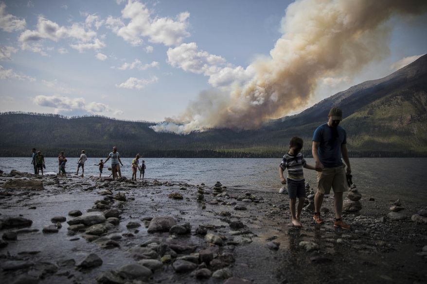 FILE - In this Sunday, Aug. 12, 2018 file photo provided by the National Park Service, people walk along the shore near Lake McDonald Lodge as the Howe Ridge Fire burns in Glacier National Park, Mont. Wildfires that have kept portions of Glacier National Park closed for two weeks are scrambling visitors’ plans and prompting some to cancel their trips. Much of Glacier’s famous Going-to-the-Sun-Road has been closed since August 12 due to a fire that’s burned more than 19 square miles (50 square kilometers). (National Park Service via AP, File)