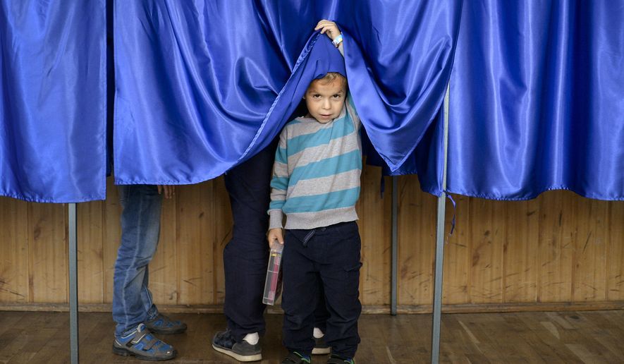 A child lifts the courtain of a voting booth as his father casts his vote, in Ciorogarla, Romania, Sunday, Oct. 7, 2018. Romanians are voting Sunday around the country for a second day on a constitutional amendment that would make it harder to legalize same-sex marriage. (AP Photo/Andreea Alexandru)