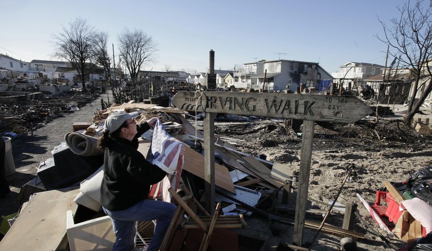 In this Nov. 14, 2012 photo, Louise McCarthy places an American flag on a street sign for Irving Walk in the Breezy Point neighborhood of Queens, N.Y. The sign survived a fire that swept through the seaside community during Superstorm Sandy two weeks earlier. Six years later, most residents have stayed. Homes have been restored, and Sandy led to a new ferry service and a rebuilt boardwalk. (AP Photo/Mark Lennihan)