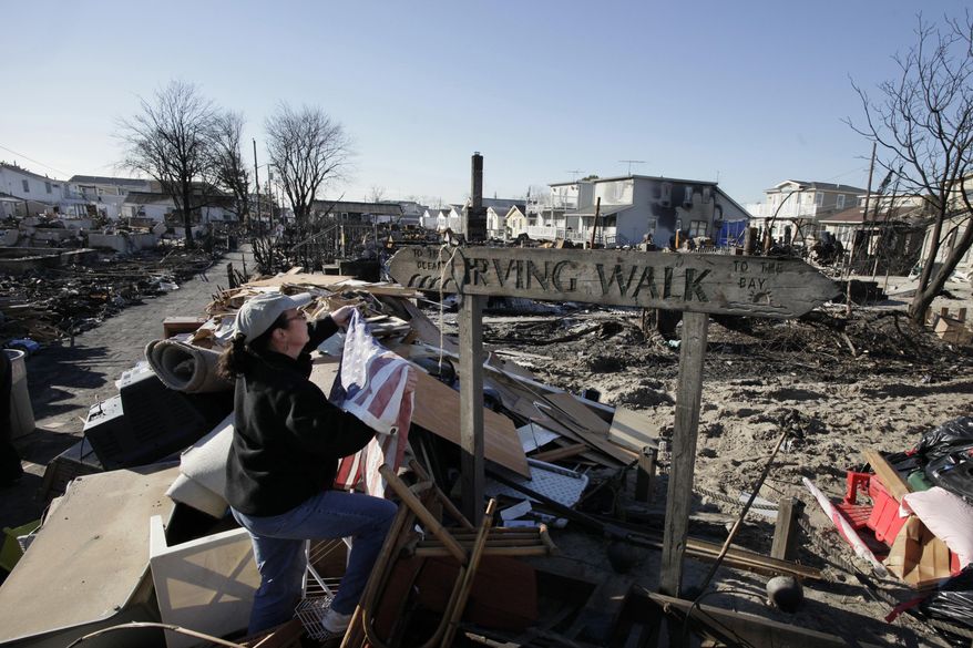 In this Nov. 14, 2012 photo, Louise McCarthy places an American flag on a street sign for Irving Walk in the Breezy Point neighborhood of Queens, N.Y. The sign survived a fire that swept through the seaside community during Superstorm Sandy two weeks earlier. Six years later, most residents have stayed. Homes have been restored, and Sandy led to a new ferry service and a rebuilt boardwalk. (AP Photo/Mark Lennihan)