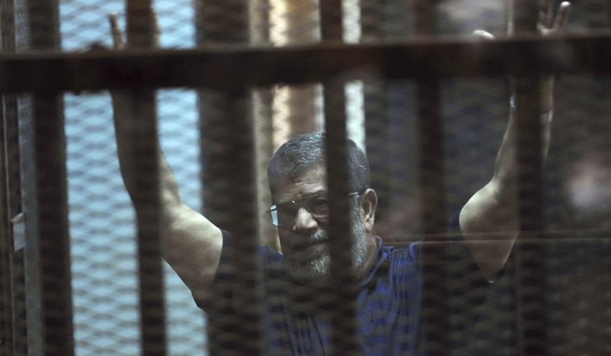 FILE - In this May 16, 2015 file photo, ousted Egyptian President Mohammed Morsi raises his hands as he sits behind glass in a courtroom, at the national police academy in a Cairo suburb, Egypt. On Wednesday, Dec. 26, 2018, two former Egyptian presidents appeared in the same Cairo courtroom. The 90-year-old Mubarak testified in a retrial of Mohammed Morsi. Mubarak whose nearly three-decade rule was ended by a popular uprising in 2011, was seen walking into the courtroom with a cane along with his two sons Alaa and Gamal. (AP Photo/Ahmed Omar, File)