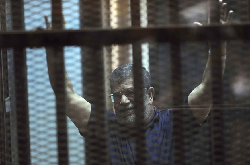 FILE - In this May 16, 2015 file photo, ousted Egyptian President Mohammed Morsi raises his hands as he sits behind glass in a courtroom, at the national police academy in a Cairo suburb, Egypt. On Wednesday, Dec. 26, 2018, two former Egyptian presidents appeared in the same Cairo courtroom. The 90-year-old Mubarak testified in a retrial of Mohammed Morsi. Mubarak whose nearly three-decade rule was ended by a popular uprising in 2011, was seen walking into the courtroom with a cane along with his two sons Alaa and Gamal. (AP Photo/Ahmed Omar, File)