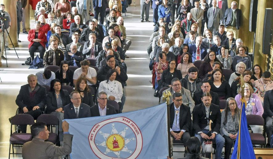 The tribal flag of the Standing Rock Sioux Nation is held by North Dakota Indian Affairs Commissioner Scott Davis, left, and tribal council member Frank White Bull Jr. as chairman Mike Faith gives remarks on Thursday, Jan. 17, 2019 in Memorial Hall of the state capitol in Bismarck, N.D. White Bull said the Lakota word for flag is "Wapaha." The flags of the Mandan, Hidatsa & Arikara, the Spirit Lake Sioux, Turtle Mountain Band of Chippewa, Sisseton-Wahpeton-Oyate as well as the Standing Rock Sioux are now on display outside the office of Governor Doug Burgum and Lt. Gov. Brent Sanford, who attended the ceremony and are seated front left. (Tom Stromme/The Bismarck Tribune via AP)