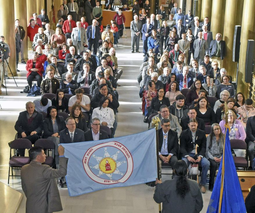The tribal flag of the Standing Rock Sioux Nation is held by North Dakota Indian Affairs Commissioner Scott Davis, left, and tribal council member Frank White Bull Jr. as chairman Mike Faith gives remarks on Thursday, Jan. 17, 2019 in Memorial Hall of the state capitol in Bismarck, N.D. White Bull said the Lakota word for flag is "Wapaha." The flags of the Mandan, Hidatsa & Arikara, the Spirit Lake Sioux, Turtle Mountain Band of Chippewa, Sisseton-Wahpeton-Oyate as well as the Standing Rock Sioux are now on display outside the office of Governor Doug Burgum and Lt. Gov. Brent Sanford, who attended the ceremony and are seated front left. (Tom Stromme/The Bismarck Tribune via AP)