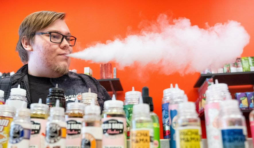FILE--In this Nov., 2018 file photo, shift manager Shaun Miller exhales vapor while working behind the vape bar at Generation V in Omaha, Neb. An apparent surge in vaping among Nebraska teenagers is prompting a new push from lawmakers to raise the age limit on e-cigarettes from 18 to 21 and ban their use in bars, restaurants and workplaces. School officials say the crackdown would help them fight the growing use of e-cigarettes among young people. (Kent Sievers/Omaha World-Herald via AP, File)