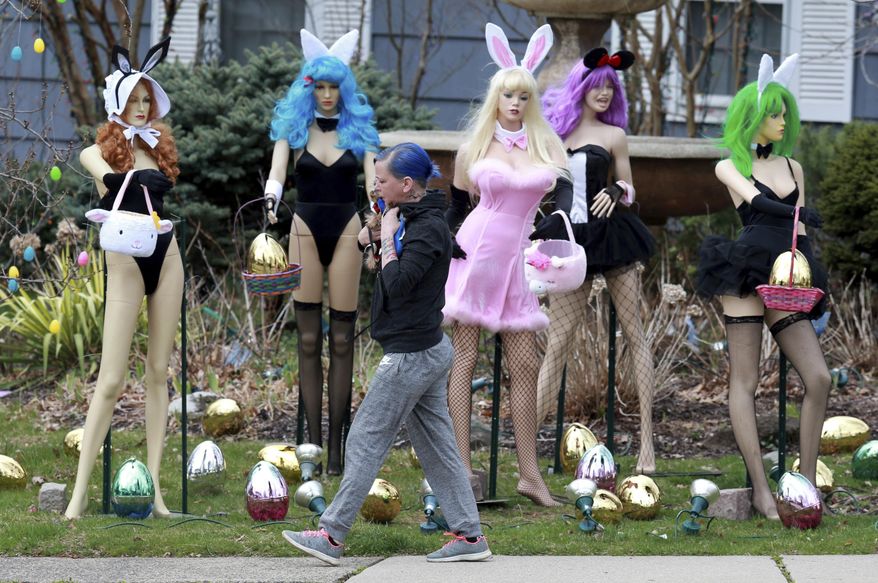 A woman walks carrying her dog past an Easter display in Clifton, N.J., Tuesday, April, 9, 2019. A New Jersey woman unhappy with her neighbor's racy Easter display used some garden shears to damage it.But the display's owner says it will soon be back up. The display at a dental office in Clifton featured five mannequins dressed in lingerie, all holding Easter baskets and surrounded by Easter eggs. It had drawn mixed reviews from neighbors, as well as passers-by who stopped to take photos. ( Ed Murray/NJ Advance Media via AP)