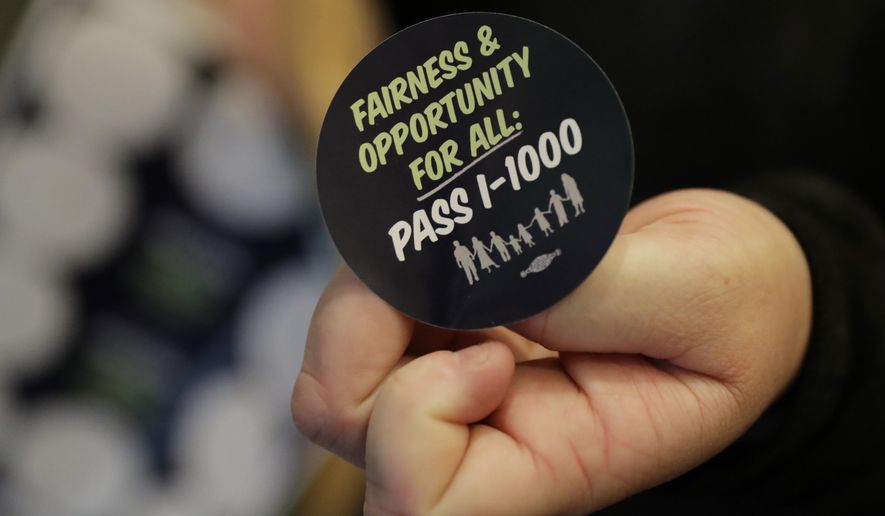 A volunteer holds a sticker showing support for Initiative 1000 outside a joint Washington state House and Senate committee, Thursday, April 18, 2019, at the Capitol in Olympia, Wash. Initiative 1000 would allow the state government to use affirmative action policies that do not constitute preferential treatment to remedy discrimination in public employment, education and contracting. (AP Photo/Ted S. Warren)