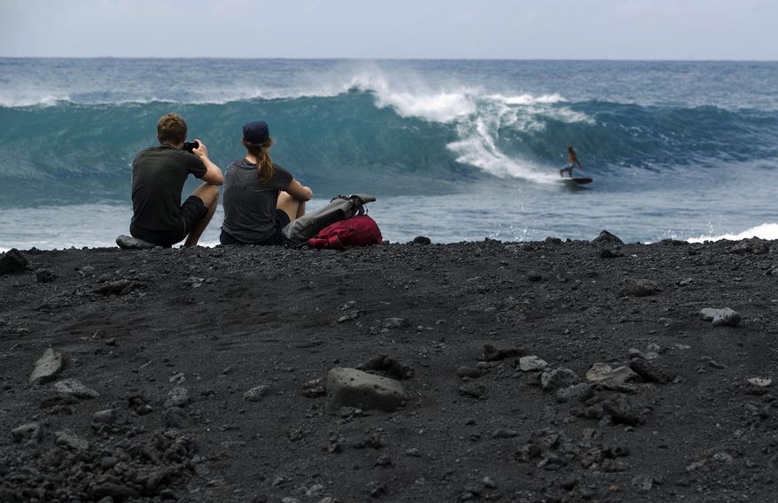 In this Tuesday, April 23, 2019 photo, visitors Bruno Hoschstrasser, left, and Mirjam Grylka, both from Switzerland, watch surfers from a newly formed black sand beach below Kilauea volcano near Kapoho, Hawaii. The beach was created as lava from the 2018 Kilauea eruption entered the ocean, broke apart and churned through the water. Before the eruption, the shoreline was about 100 yards inland from where it is now. (AP Photo/Marco Garcia)