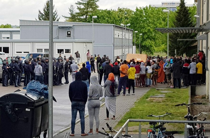 German police officers and migrants stand in front of an asylum center after a woman was found dead in the center in Regensburg, Germany, Saturday, May 11, 2019. German police say it took them several hours to recover the body of the 31-year-old African woman from the center because angry residents there pelted officers with stones and other objects. (Alexander Auer/dpa via AP)