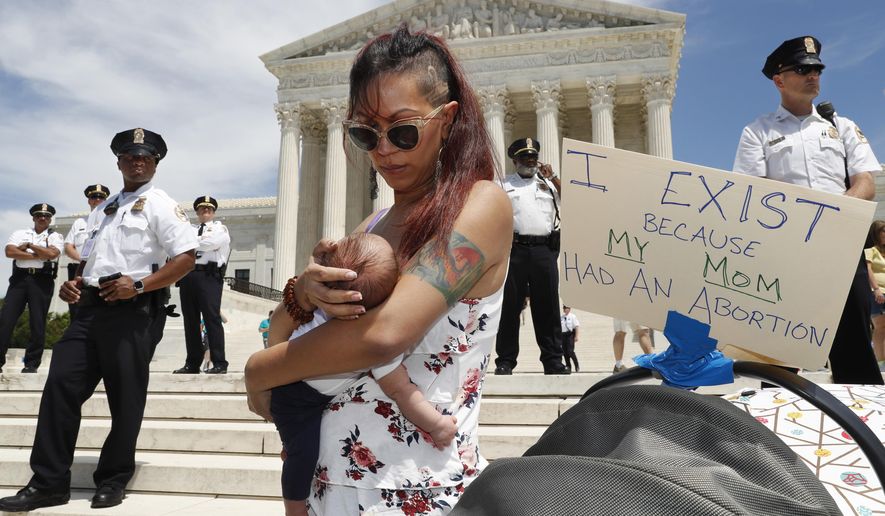 As security guards the steps of the Supreme Court, Kristin Mink of Silver Spring, Md., holds her three-week-old daughter by a sign she brought that says, "I exist because my Mom had an abortion," as Mink joined a protest against abortion bans, Tuesday, May 21, 2019, outside the Supreme Court in Washington. Being able to access abortion services in an earlier pregnancy made it possible for Mink to now give birth to this child. (AP Photo/Jacquelyn Martin)
