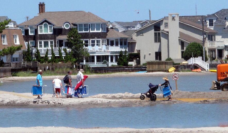 In this July 31, 2017 photo beachgoers cross over one of numerous large pools of water that formed on the beach in Margate N.J., due to heavy rains. The water was blocked from draining into the ocean by new sand dunes being built as part of a storm protection program that Margate residents vigorously fought, claiming that the dunes would cause exactly the type of standing water that occurred. Now some residents are pushing for construction of a boardwalk to mitigate what they say is damage to the beach caused by the dune project. (AP Photo/Wayne Parry)