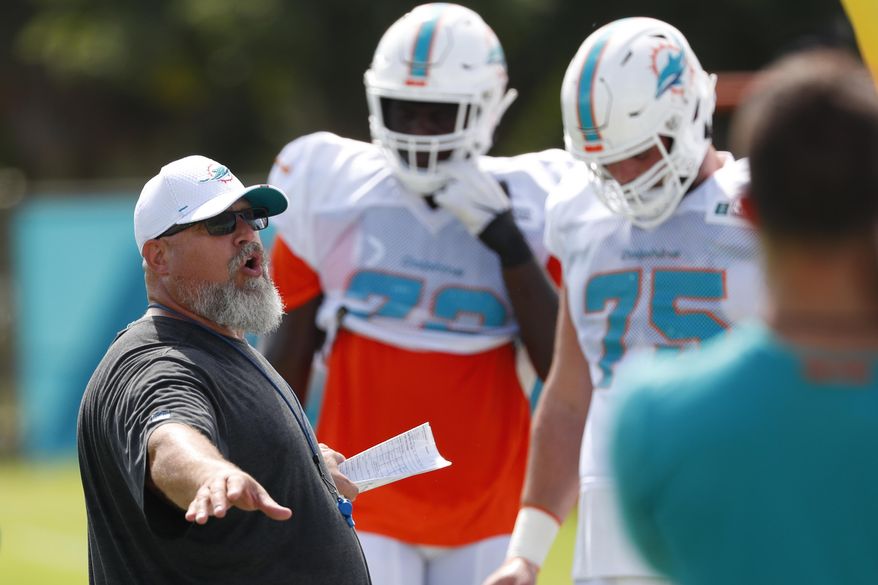 FILE - In this July 30, 2019, file photo, Miami Dolphins offensive line coach Dave DeGuglielmo, left, runs players through drills at the teams NFL football training camp, in Davie, Fla. As part of rookie coach Brian Flores’ staff, offensive line coach Dave DeGuglielmo is trying to clean up a mess for the Miami Dolphins, which is nothing new. DeGuglielmo also was a Dolphins assistant in 2009-11, and for the last part of 2017. “I really like it here,” he said Tuesday, Sept. 3, 2019. “I’m like a stray dog _ they keep feeding me, I keep coming back.”(AP Photo/Wilfredo Lee, File)