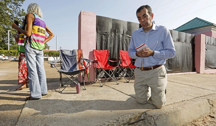 Dani Choufani recites the rosary outside the Jackson Women's Health Organization clinic, while clinic escorts wait for incoming patients in Jackson, Miss., Wednesday, Oct. 2, 2019. The Jackson City Council voted 3-1 Tuesday to enact a local law limiting amplified sound outside health care facilities and creating buffer zones to move protesters further from the entrances. The law is set to take effect in about a month, and opponents say it unconstitutionally limits their right to free speech. A court challenge is likely. (AP Photo/Rogelio V. Solis)