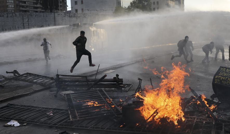 In this Oct. 29, 2019, file photo, anti-government protesters run from police spraying water cannons where a street barricade burns, set by demonstrators, in Santiago, Chile. From Honduras to Chile, popular frustration with anemic economic growth, entrenched corruption and gaping inequality is driving the region’s middle classes to rebel against incumbents of all ideological bents in what has been dubbed by some the Latin American Spring. (AP Photo/Rodrigo Abd, File)
