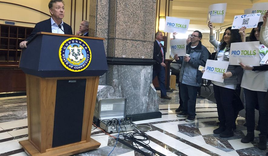 Connecticut Gov. Ned Lamont, left, answers questions from the media following a meeting of the State Bond Commission, Wednesday, Dec. 18, 2019, at the Legislative Office Building in Hartford, Conn. A group of mostly Fairfield County residents hold signs, right, showing their support for tolls. The contentious issue of how to fund transportation improvements in the state is expected to be taken up in a special session of the General Assembly in January. (AP Photo/Susan Haigh)
