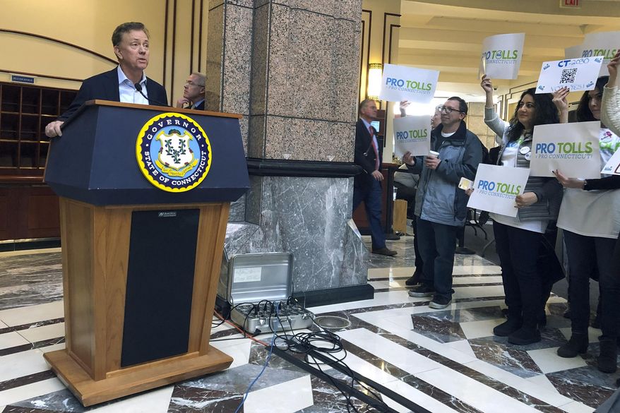 Connecticut Gov. Ned Lamont, left, answers questions from the media following a meeting of the State Bond Commission, Wednesday, Dec. 18, 2019, at the Legislative Office Building in Hartford, Conn. A group of mostly Fairfield County residents hold signs, right, showing their support for tolls. The contentious issue of how to fund transportation improvements in the state is expected to be taken up in a special session of the General Assembly in January. (AP Photo/Susan Haigh)