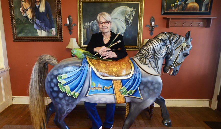 In this photo taken Dec. 3, 2019, artist Linda Luster stands in Rocky Ridge, Md., behind the carousel horse she recently painted. Some of her paintings hang on the wall. (Sam Yu/The Frederick News-Post via AP)