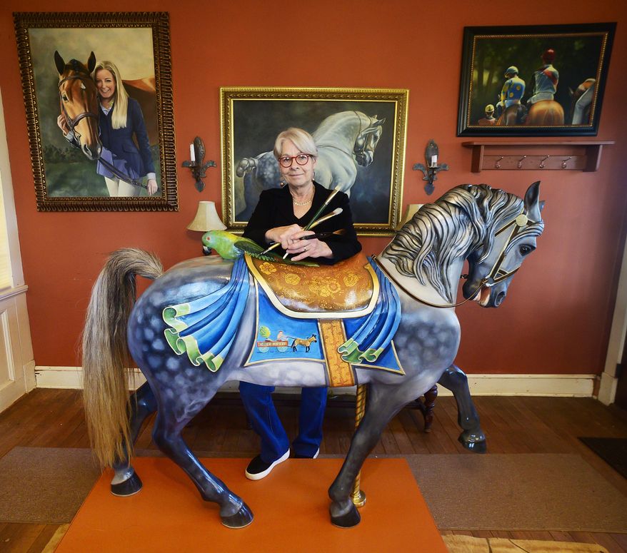In this photo taken Dec. 3, 2019, artist Linda Luster stands in Rocky Ridge, Md., behind the carousel horse she recently painted. Some of her paintings hang on the wall. (Sam Yu/The Frederick News-Post via AP)