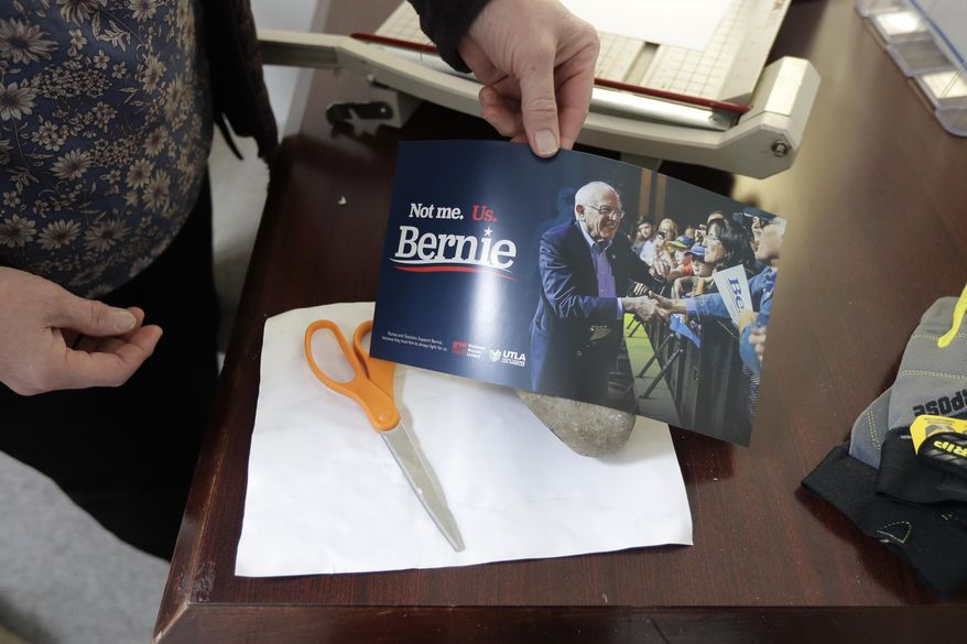 Carin Chase, director of Washington state for the campaign of Democratic presidential candidate Sen. Bernie Sanders, I-Vt., uses a Sanders campaign sign to cover a rock Saturday, Feb. 22, 2020, in Seattle that was used to break the glass of the door to Sanders' Seattle headquarters Friday night as she makes a humorous reference to a "rock-paper-scissors" game. Police are investigating, but no one was hurt in the vandalism incident, and campaign workers swept up the pieces and patched the door with plywood. (AP Photo/Ted S. Warren)